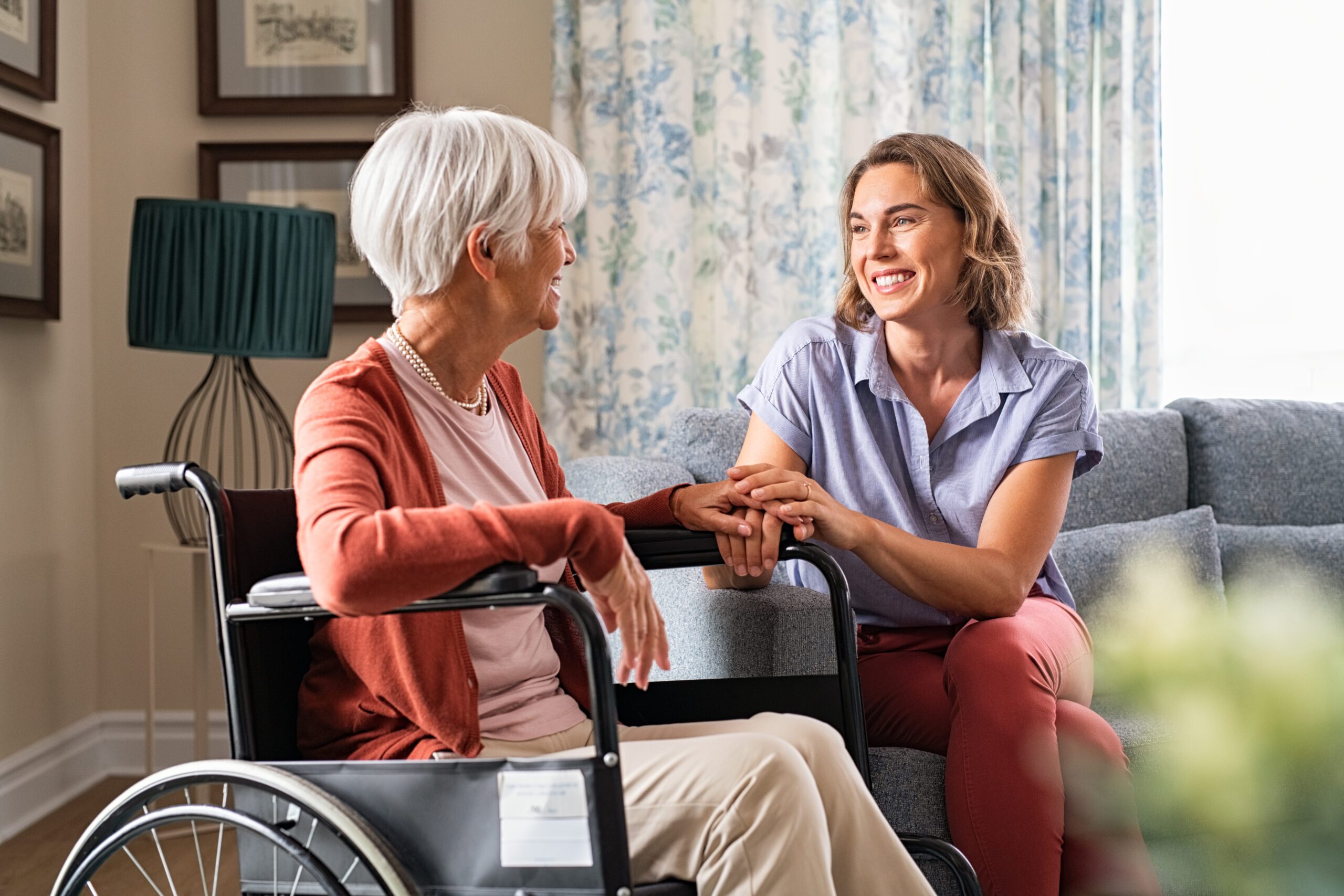 Two women sitting in a living room are chatting while holding hands, and the older of the two women is sitting in a wheelchair.